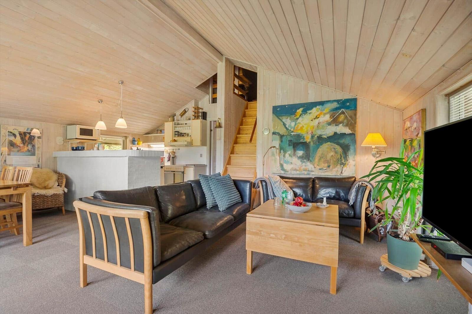 Wohnzimmer mit Sofa, Couchtisch und Fernseher. Hintergrund: Küche und Treppe.