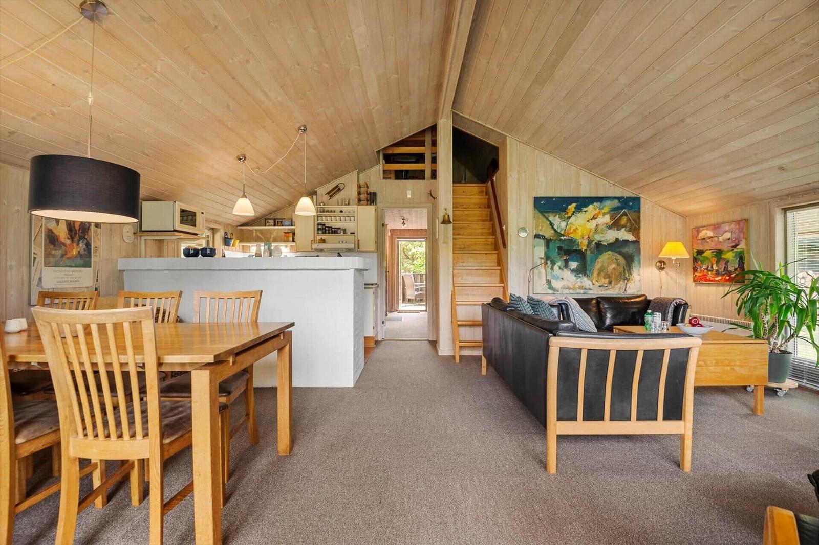 Wohn- und Essbereich mit Holzdecke, Sofa, Tisch und Stühle. Treppenhaus und Küche im Hintergrund.