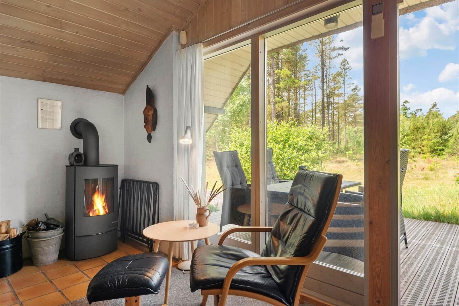 Raum mit Holzdecke, Kamin und Sitzgelegenheit. Durchgehende Glasfront zur Terrasse mit Blick auf Wald.