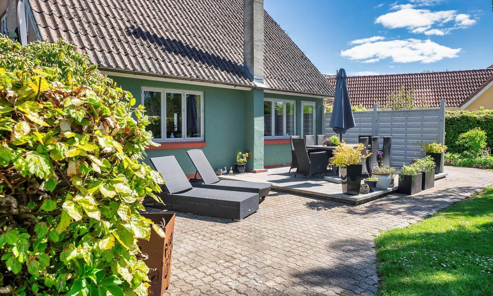 Terrace with loungers, table, and chairs. House with green paint and roof. Garden with lawn and plants.