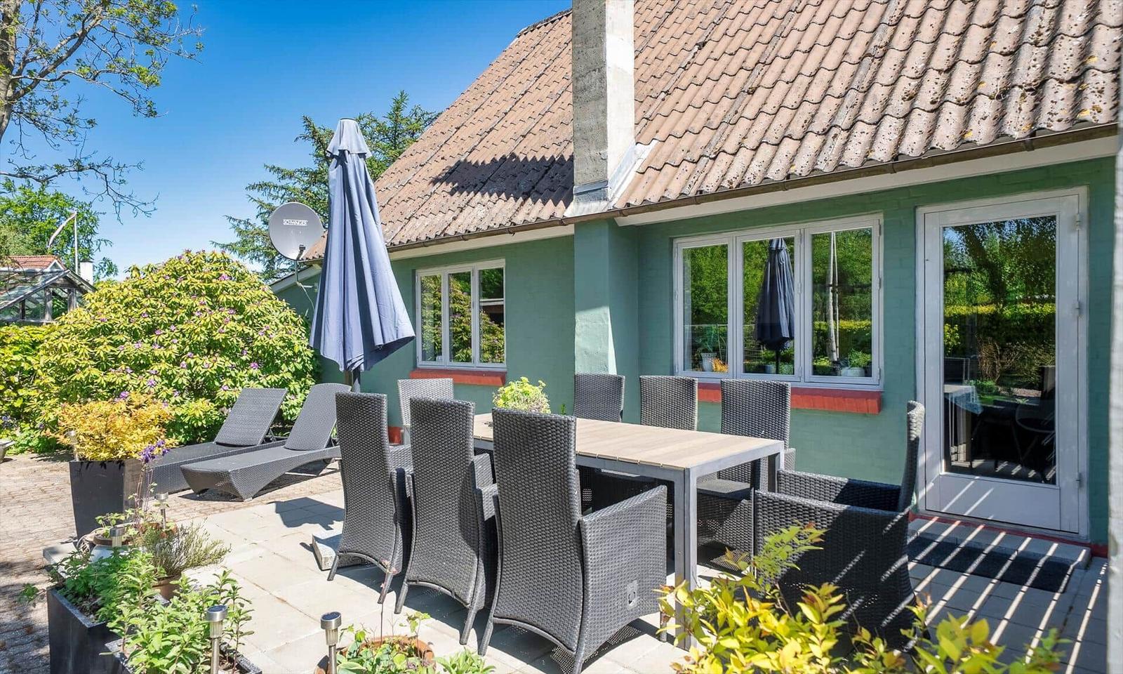 Terrace with table, chairs, and umbrella. House with green paint and tile roof.