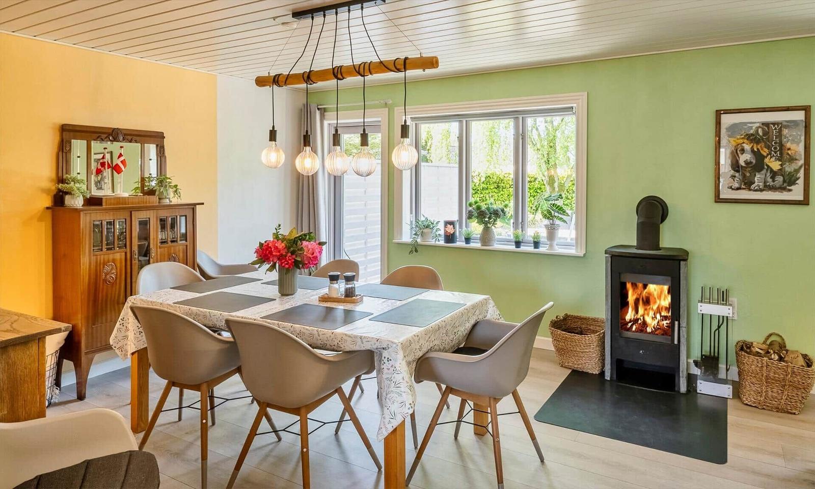 Dining room with table, chairs, and wood stove. Windows with plants and wall art.
