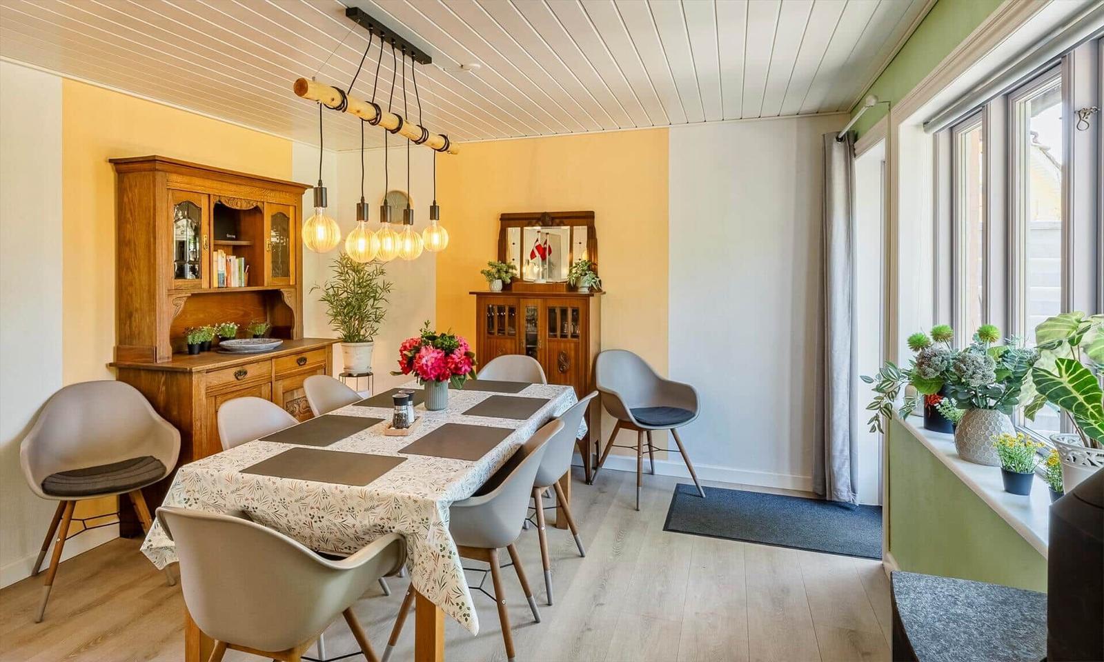 Dining room with wooden table, modern chairs, and wooden cabinet. Window sill with plants and warm lighting.