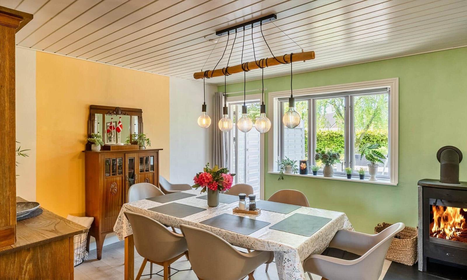 Dining room with table, chairs, and fireplace. Window with plants. Ceiling light with bulbs.