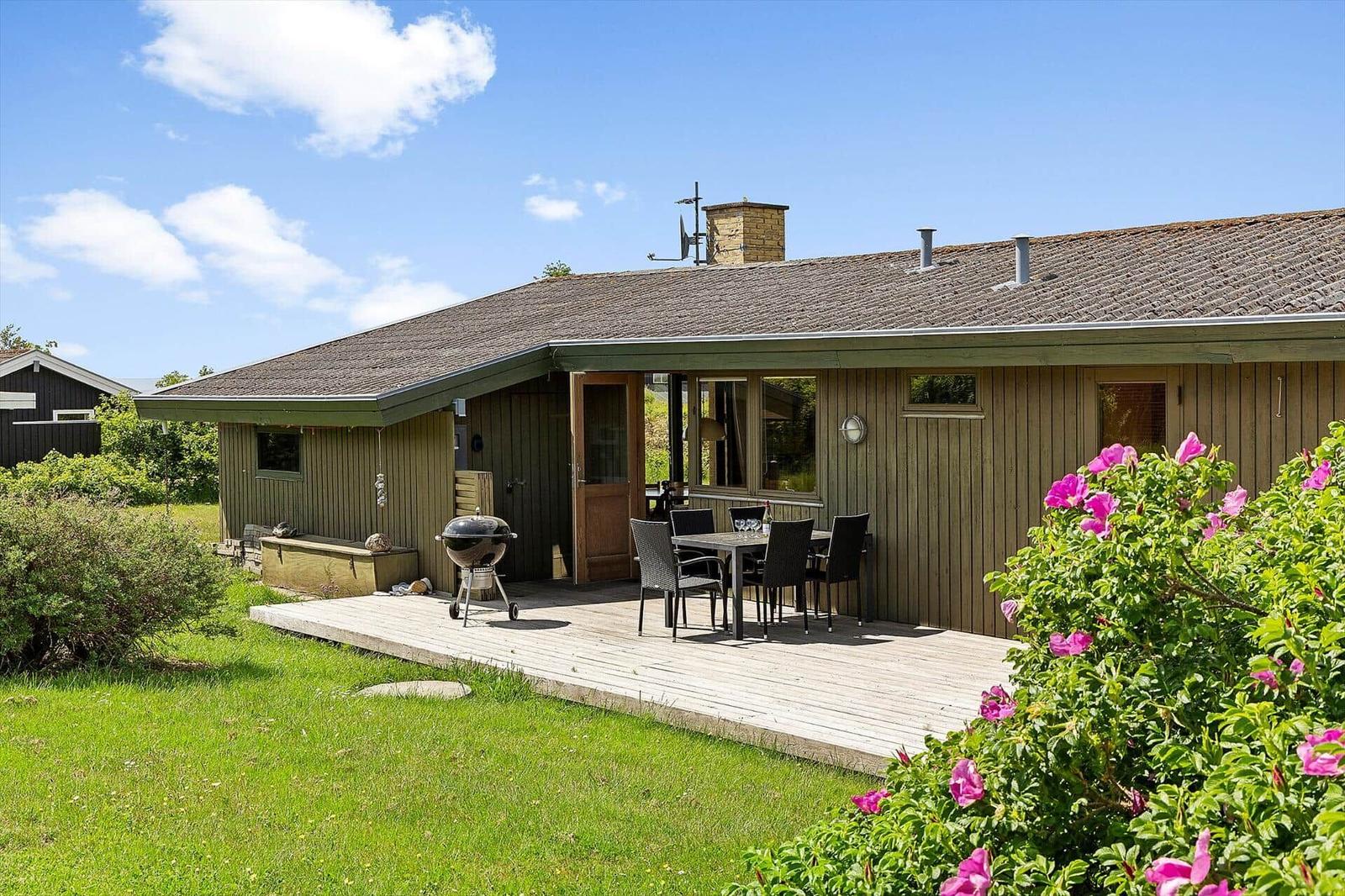 Wooden house with terrace, grill, and seating. Blooming bushes in the foreground.