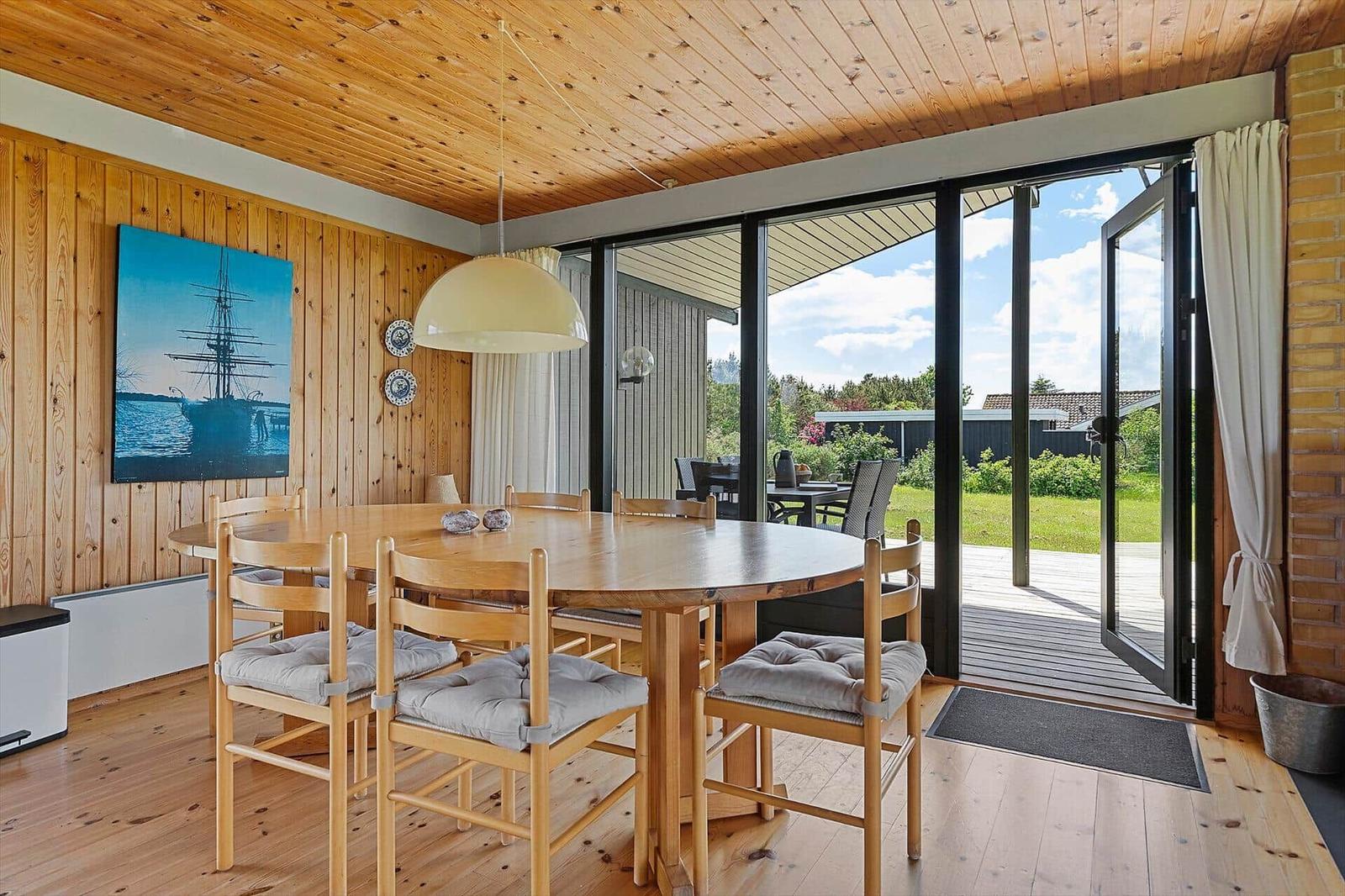 Dining area with wooden table and chairs, large window to garden, wall painting with sailboat.