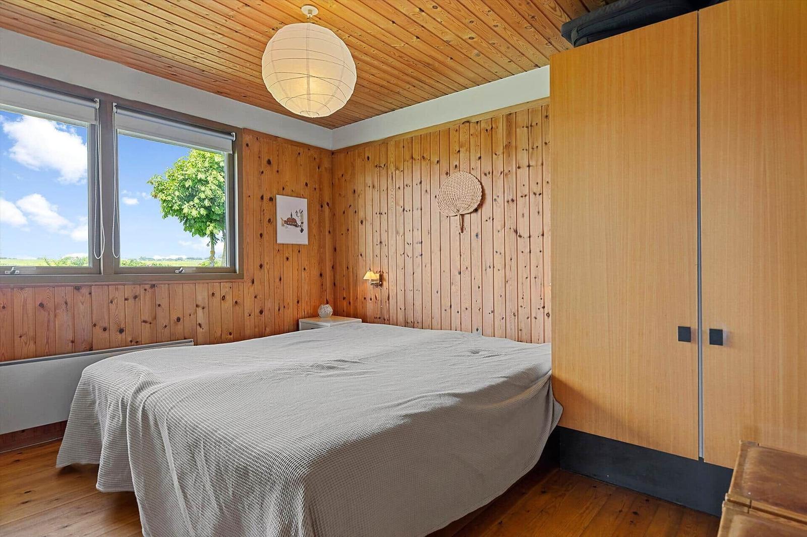 Bedroom with wooden walls, large window, and bed. Wooden ceiling, wall lamp, and wardrobe.