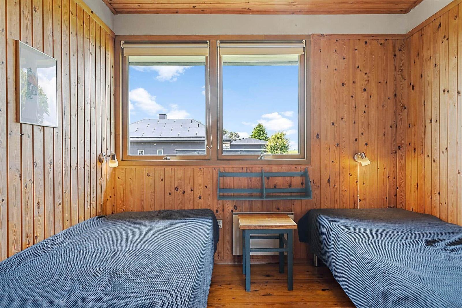 Two beds with blue bedspreads, wooden walls, and windows with a view of roofs and trees.