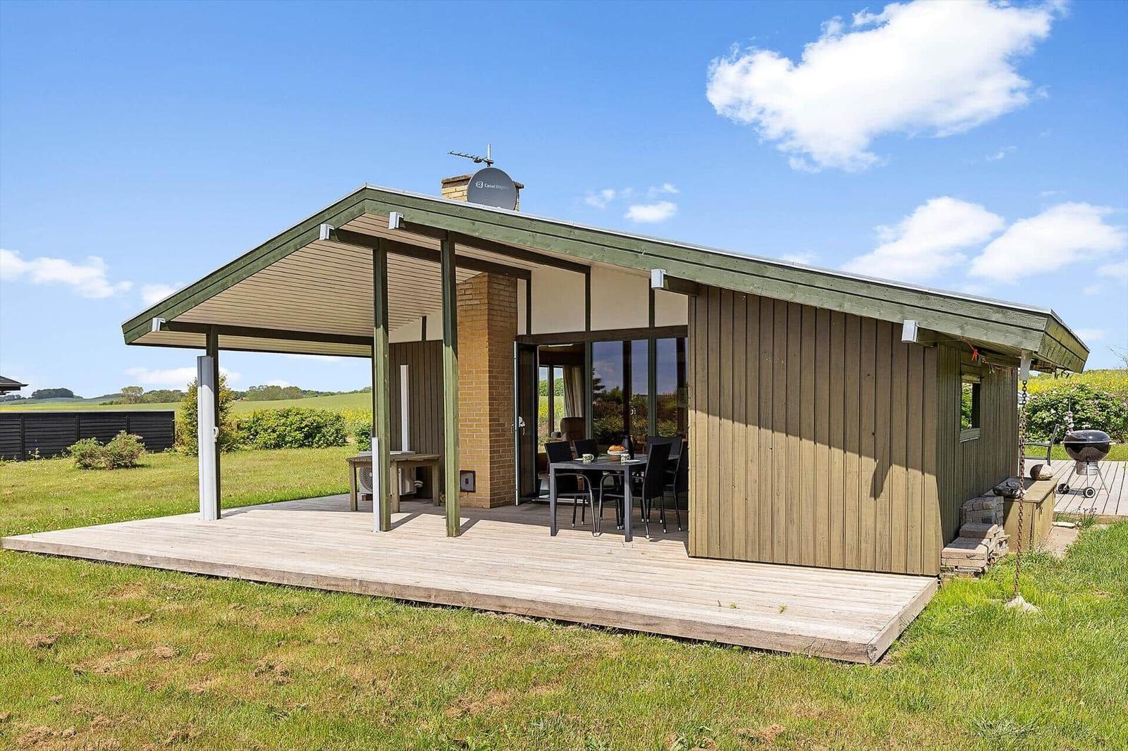 Modern wooden cabin with terrace, grill, and view of fields under blue sky.