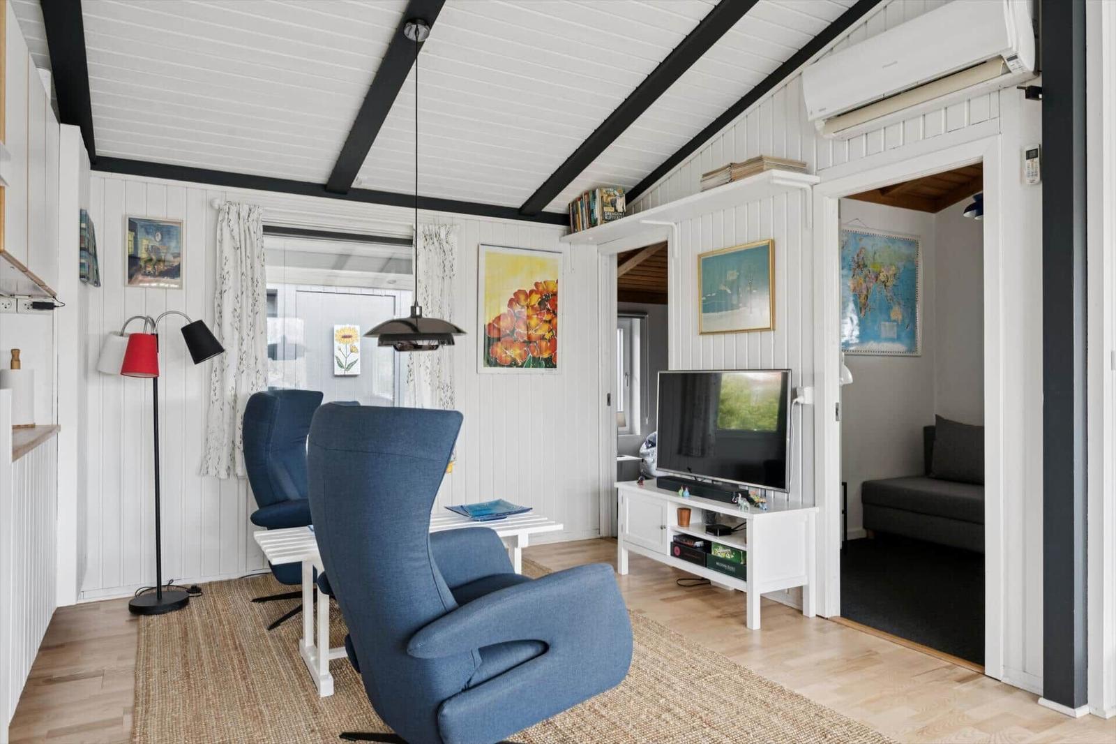 Wohnzimmer mit blauen Sesseln, Fernseher und weißen Wänden. Durchgang zu Schlafzimmer.