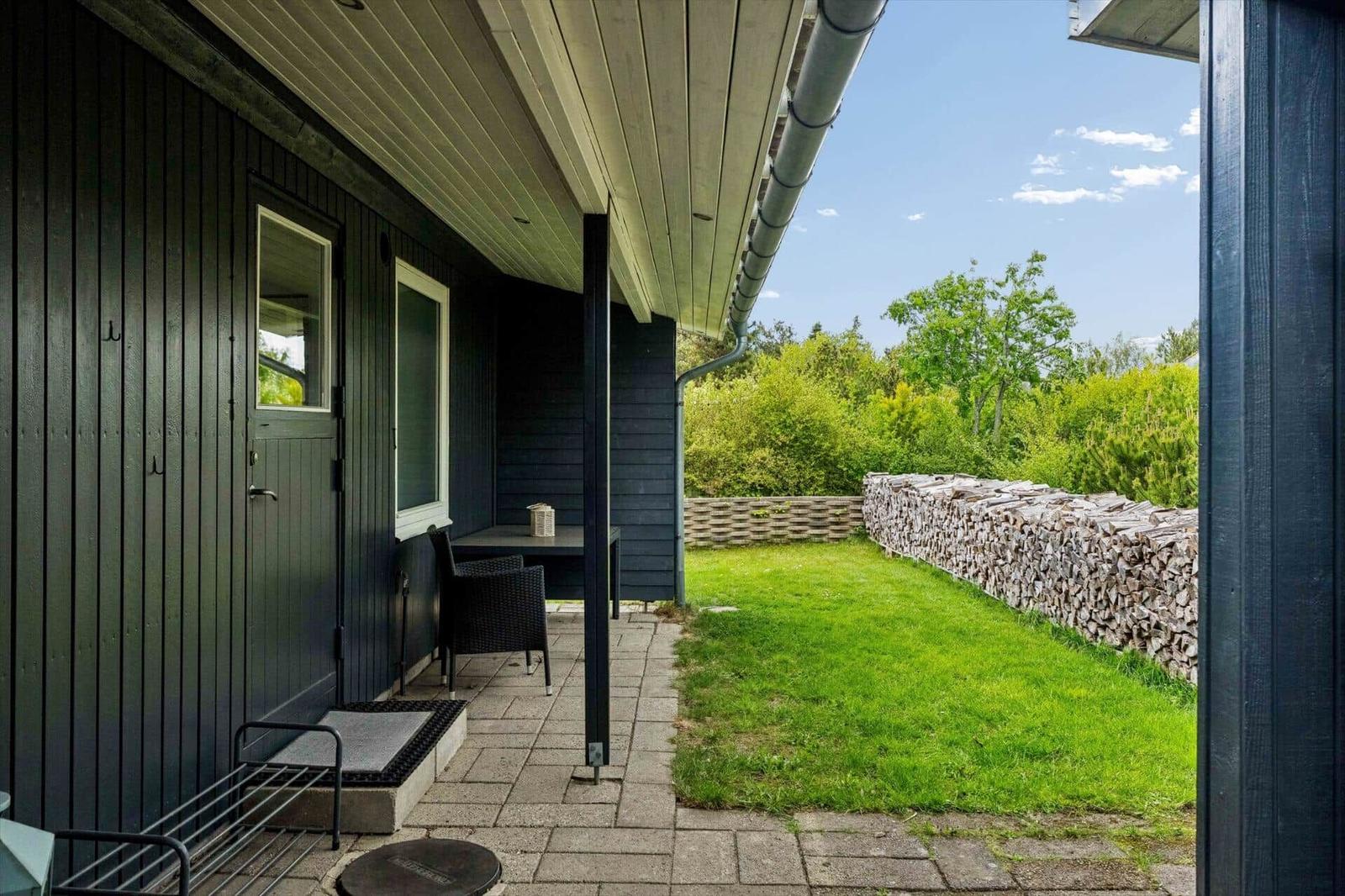 Terrasse mit Tisch und Stuhl neben grünem Garten und Holzstapel.