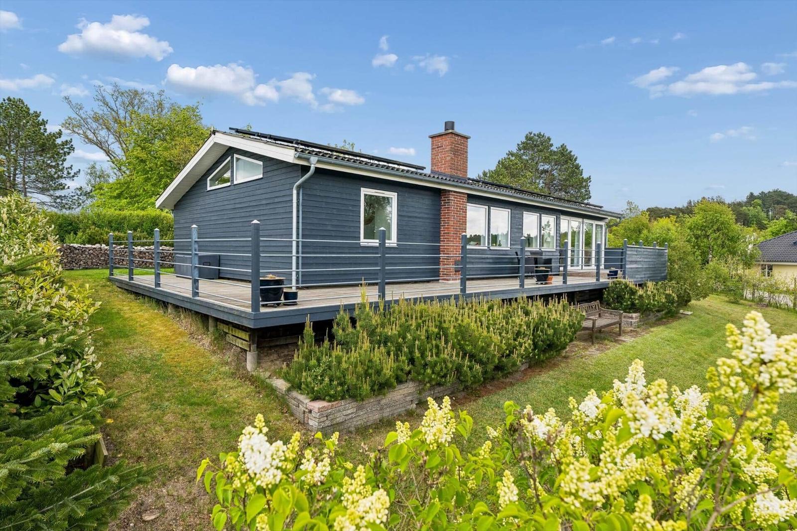 Ein Haus mit Holzterrasse und Garten. Im Hintergrund sind Bäume und ein klarer Himmel zu sehen.
