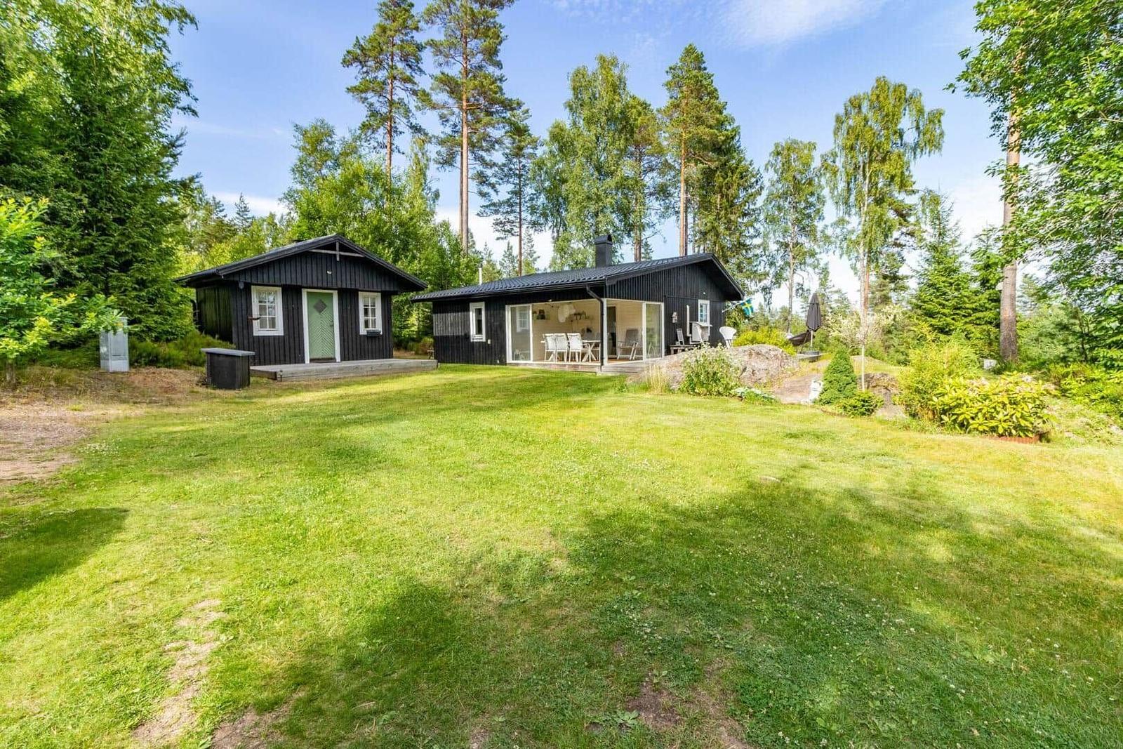 Zwei schwarze Holzhäuser mit Terrasse und Garten inmitten von Bäumen.
