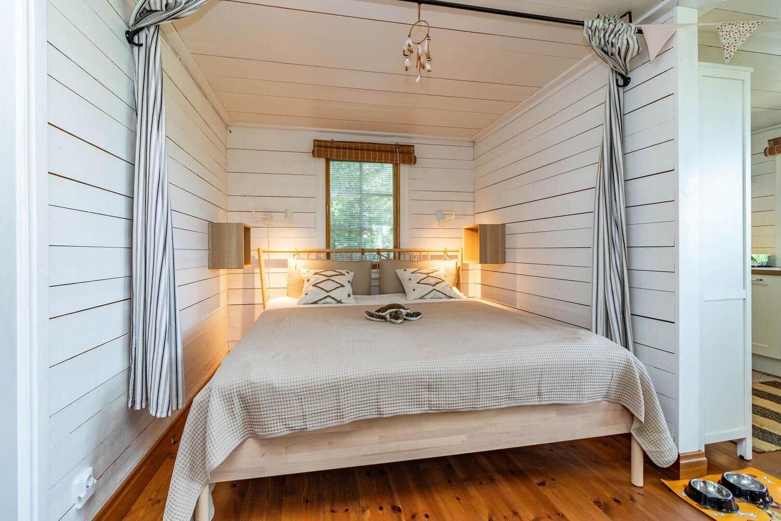 Schlafzimmer mit Bett, Holzboden und weißen Wänden. Fenster mit Rollladen und Wandlampen.