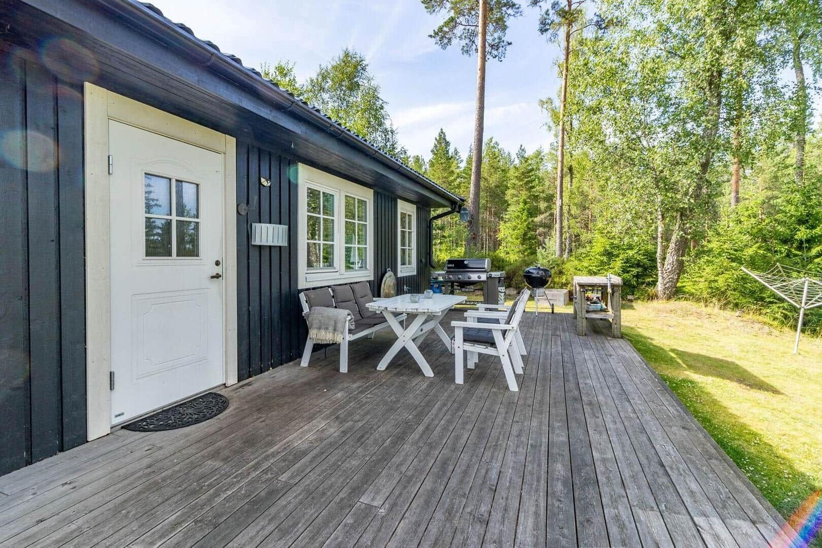 Holzterrasse mit Tisch, Stühlen und Grill neben einem Haus mit dunkler Fassade und weißer Tür.