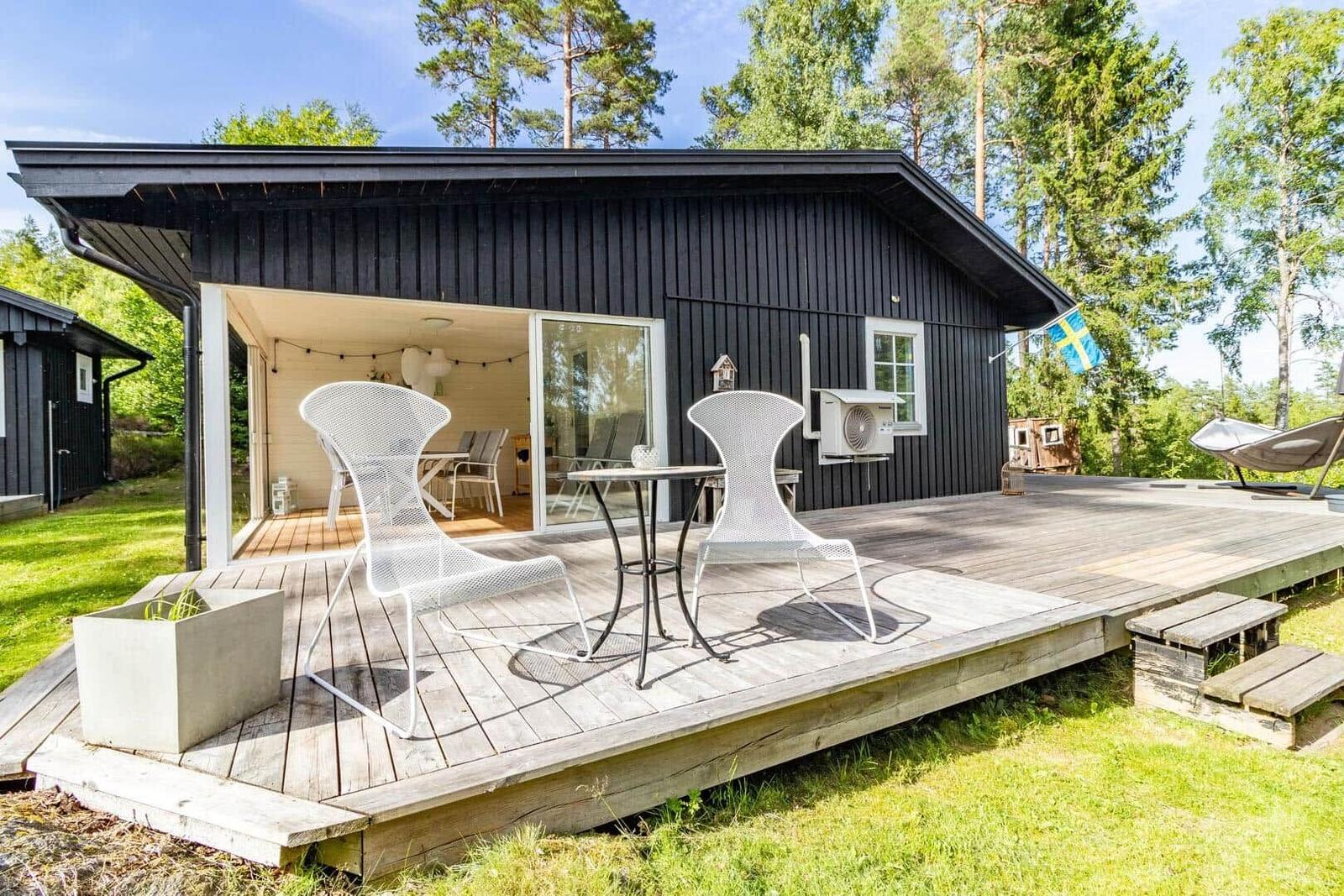 Schwarzes Holzhaus mit Terrasse, Tisch, Stühlen und schwedischer Flagge.