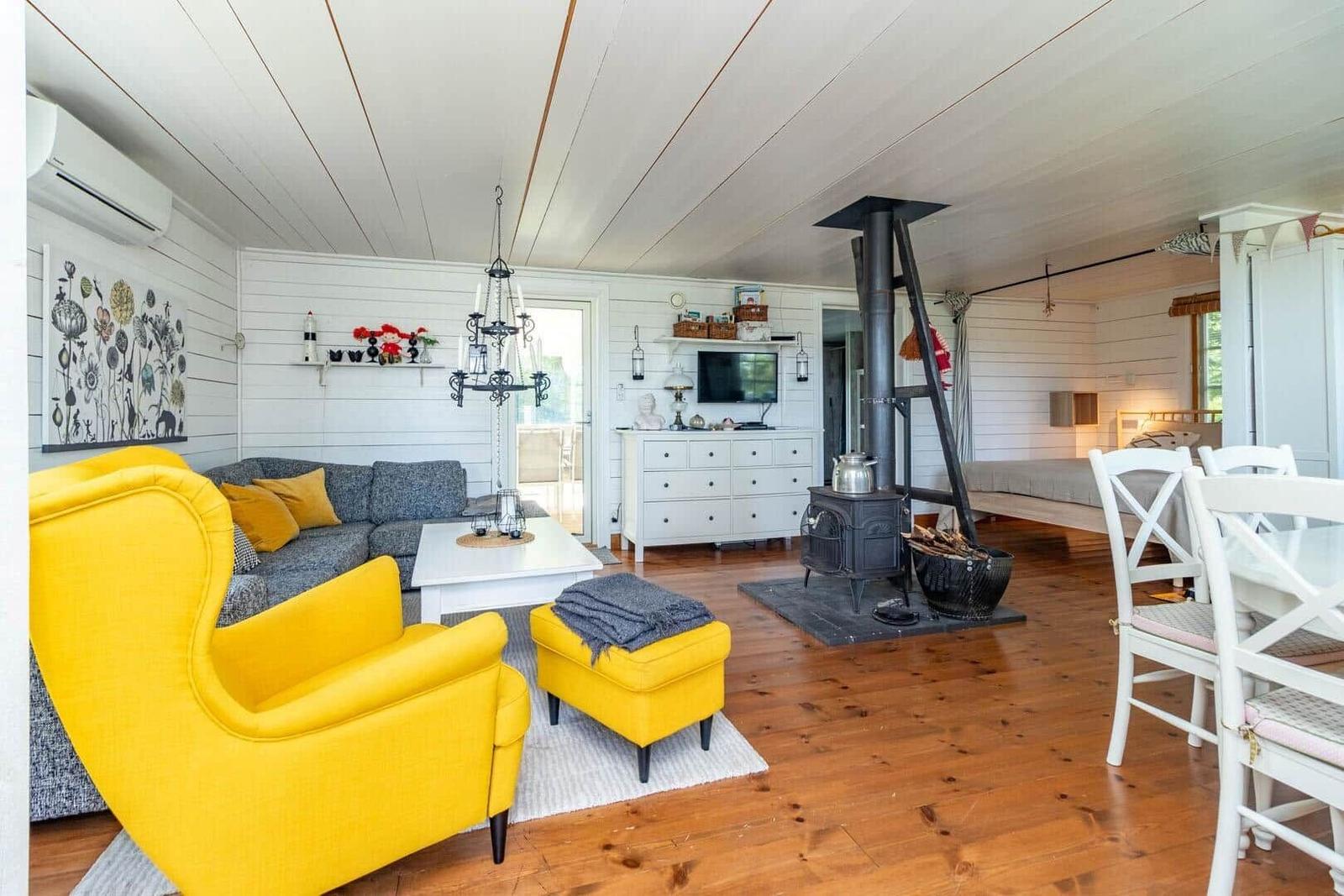 Wohnzimmer mit gelben Sesseln, Holzboden und Kaminofen. Fernseher und Essbereich sind sichtbar.