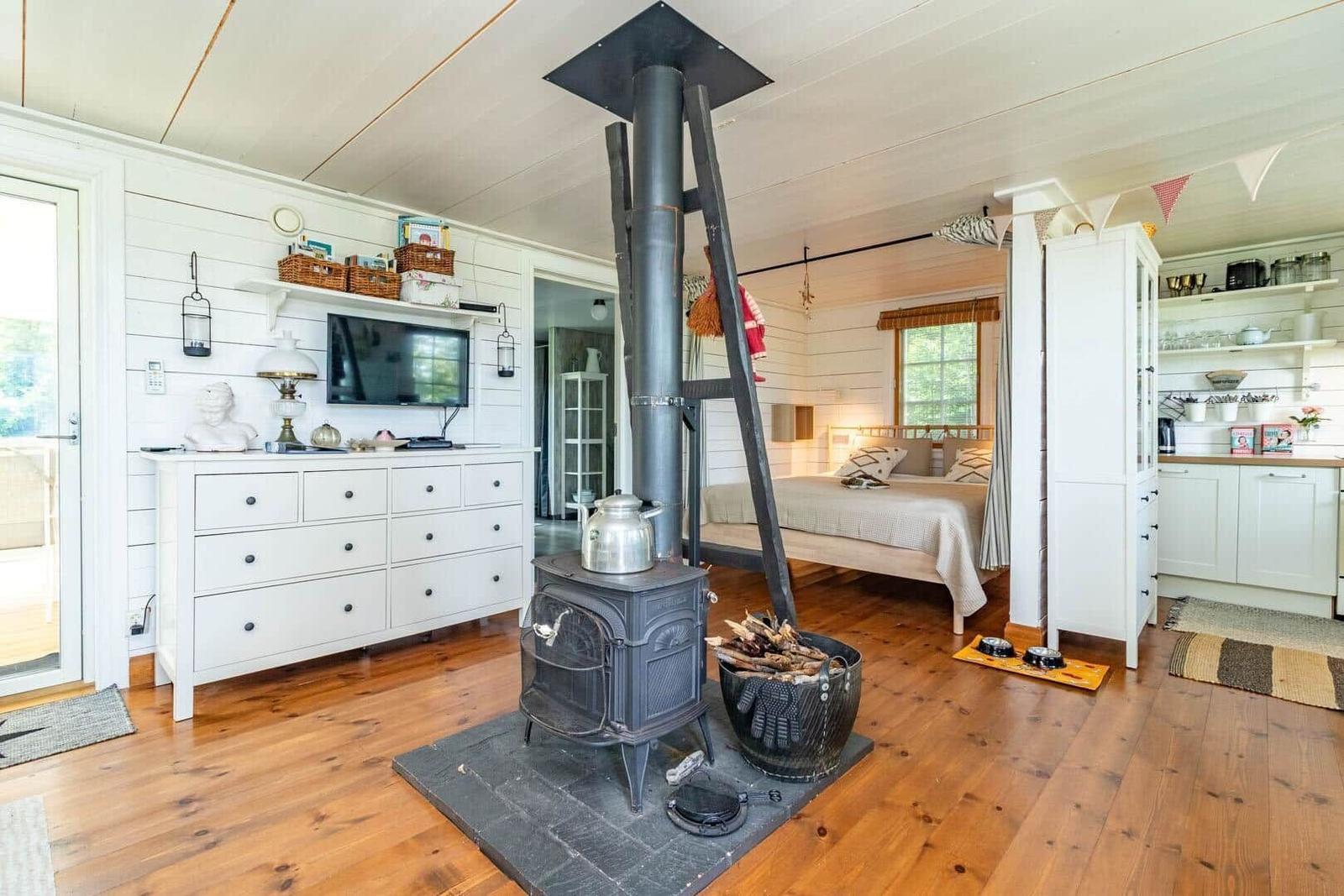Zimmer mit Kaminofen, Bett und Küchenbereich. Holzboden und weiße Wände.