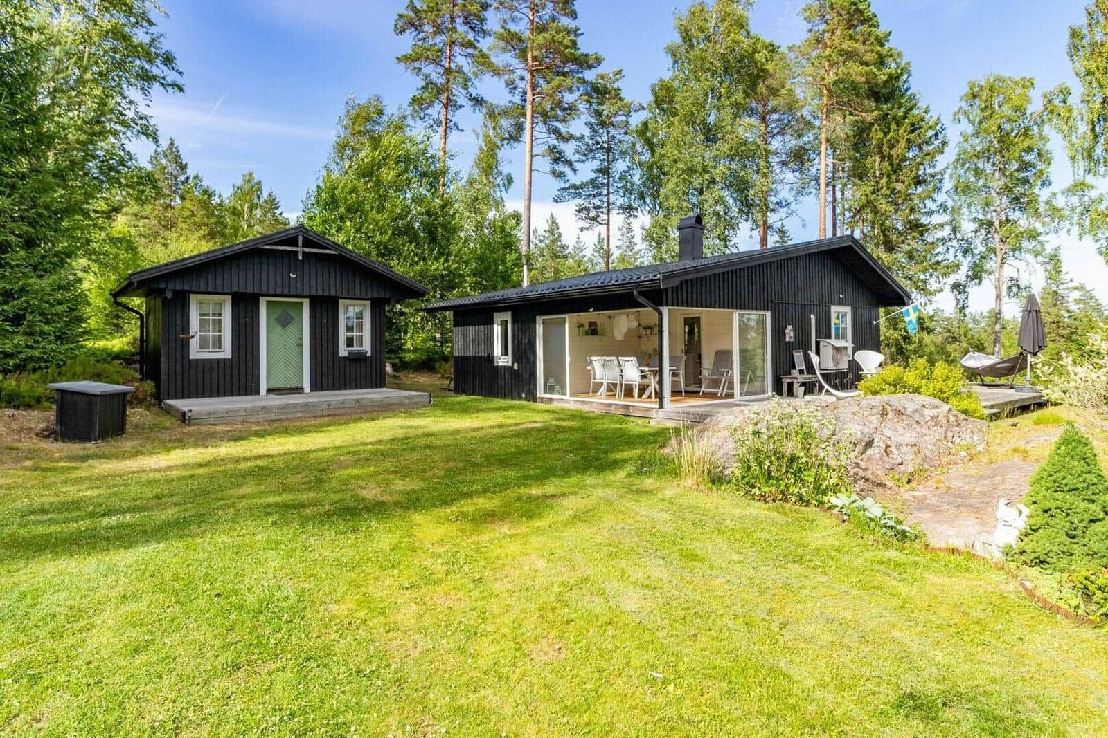 Zwei schwarze Holzhäuser mit Terrasse und Garten in einem Waldgebiet.