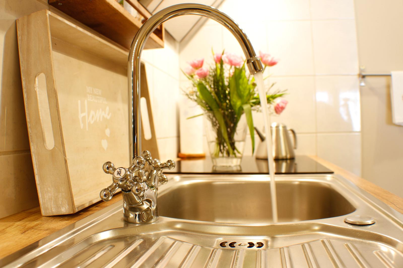 Küchenarmatur mit laufendem Wasser, Holzbrett und Blumen im Hintergrund.