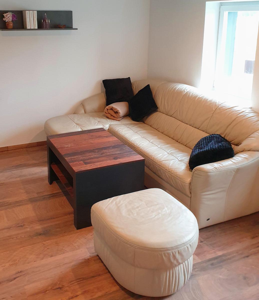 Ein Wohnzimmer mit cremefarbenem Sofa, Holztisch und Fußhocker. Wandregal mit Dekoration.