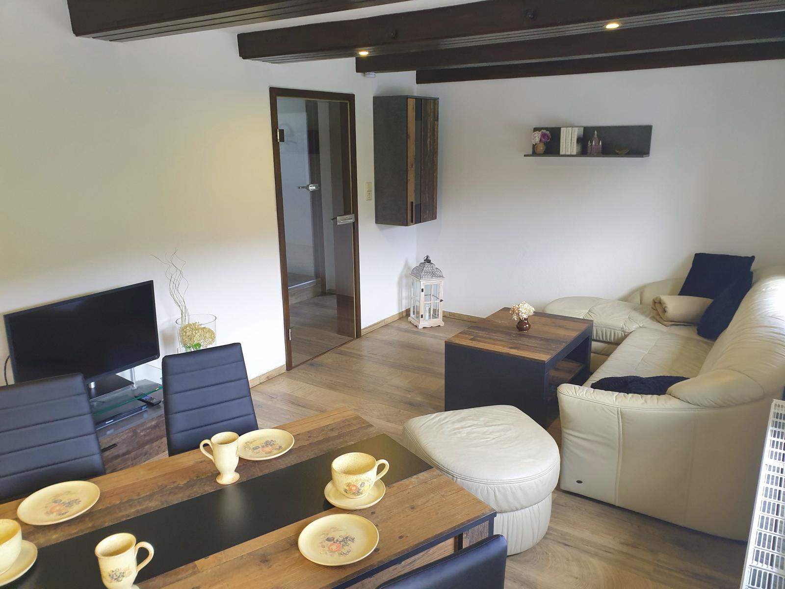 Wohn- und Essbereich mit Sofa, Tisch und Fernseher. Holzboden und Deckenbalken.