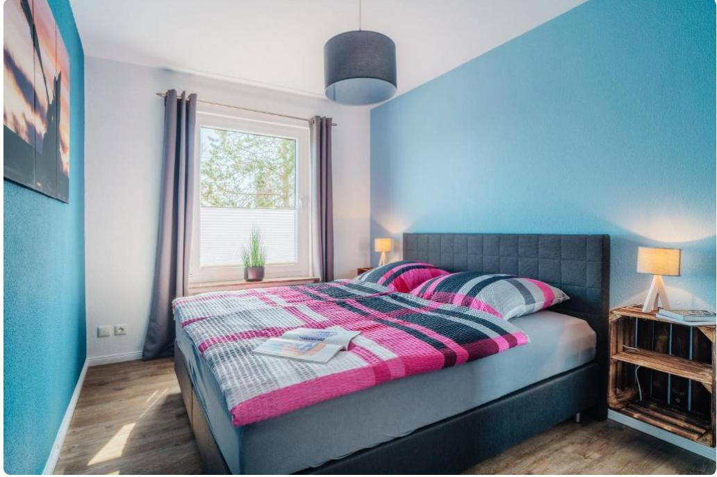 Schlafzimmer mit Bett, Fenster und blauen Wänden. Neben dem Bett steht ein Nachttisch.