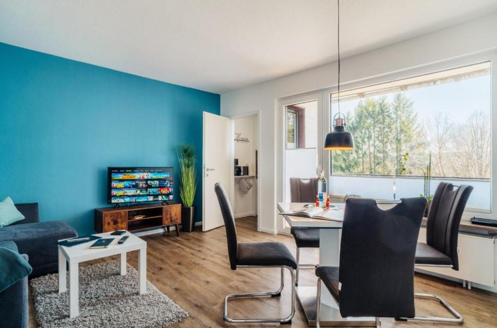 Wohn- und Essbereich mit TV, Tisch und Stühlen. Blaue Wand, Holzboden und Fenster mit Blick.