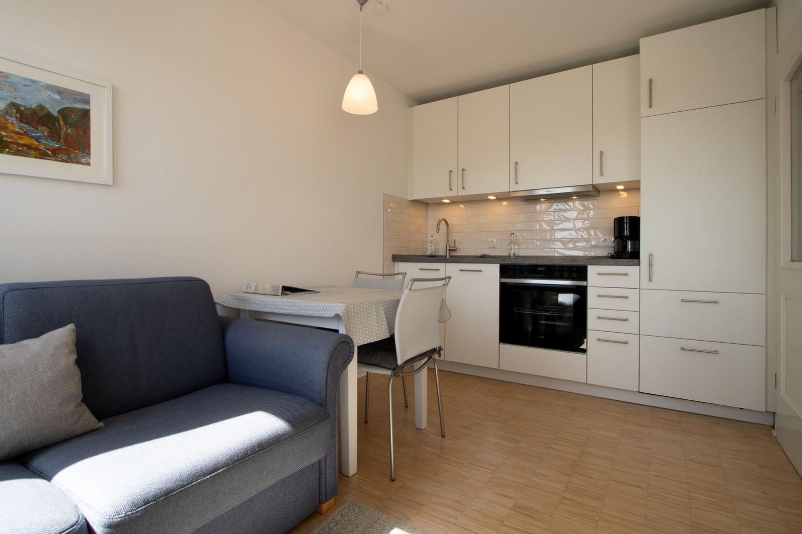 Living and kitchen area with sofa, table, white cabinets, and wooden floor.