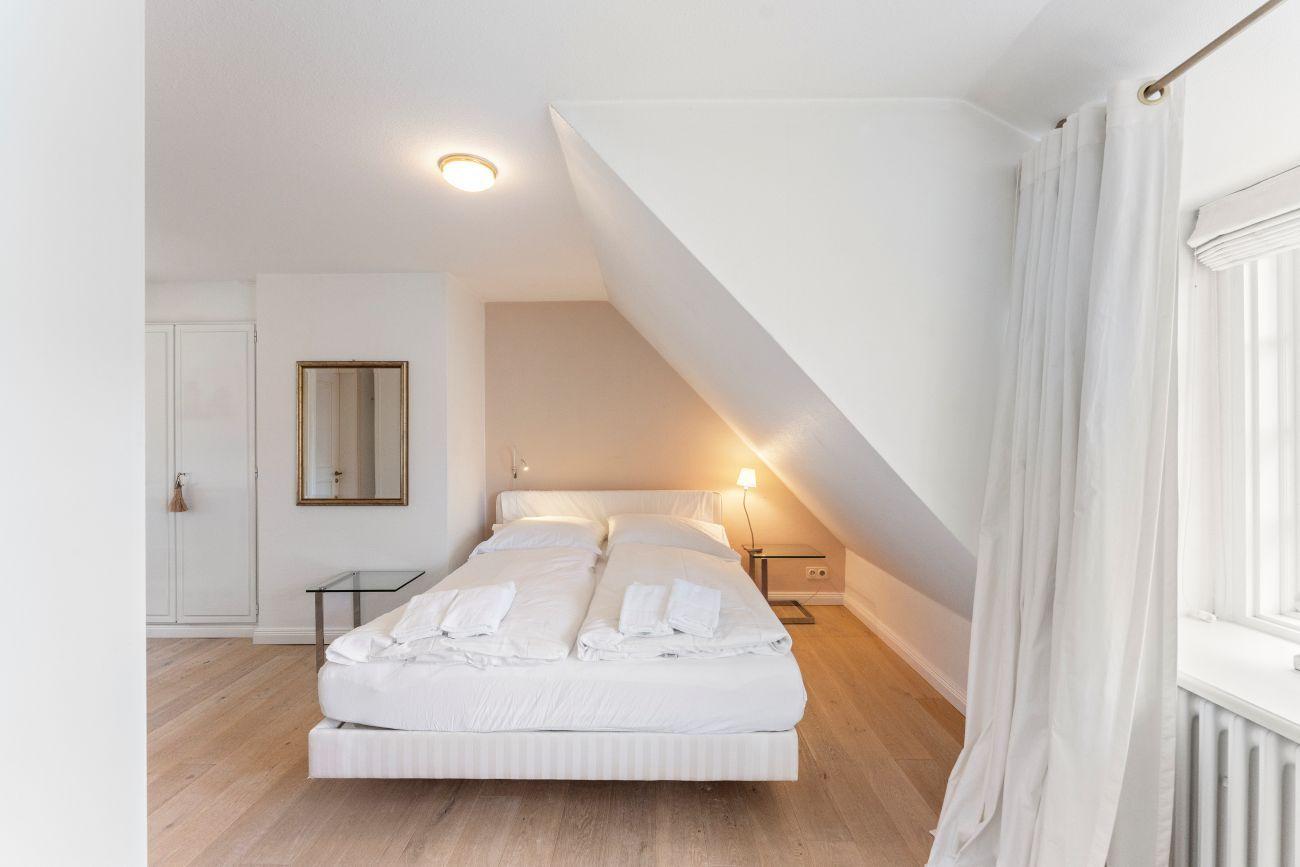 Schlafzimmer mit Doppelbett, Holzboden und schräger Decke. Fenster mit weißen Vorhängen.