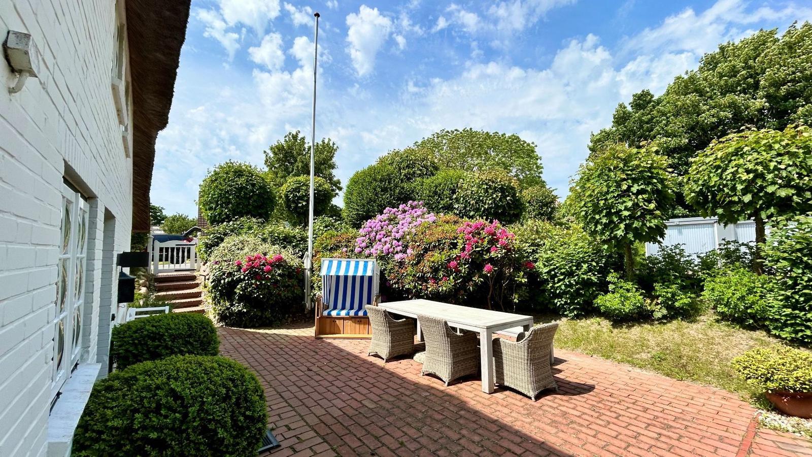 Terrasse mit Tisch, Stühlen und Strandkorb. Umgeben von Blumen und Bäumen. Hintergrund: weißes Haus mit Schopf.