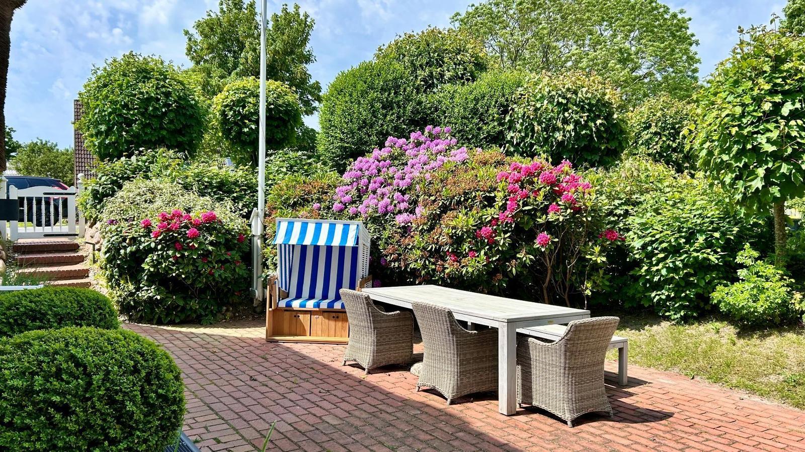 Terrasse mit Tisch, Stühlen und Strandkorb umgeben von Blumen und Bäumen.