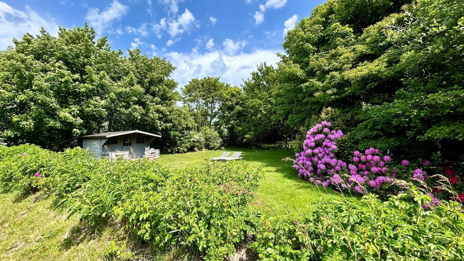 Grüner Garten mit Hütte, Sonnenliegen und lila Blumen unter blauem Himmel.