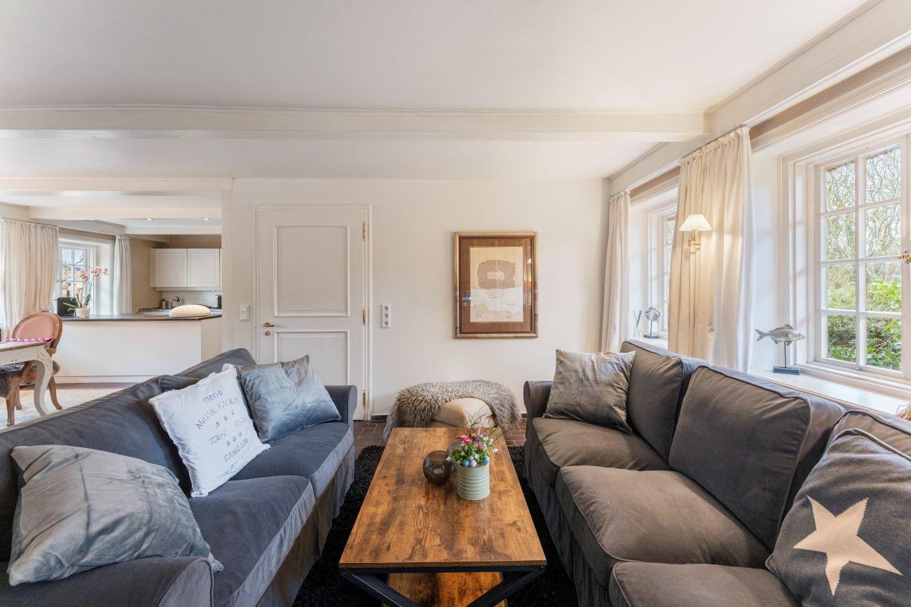 Wohnzimmer mit zwei Sofas, Holztisch und Fenstern. Küche im Hintergrund sichtbar.