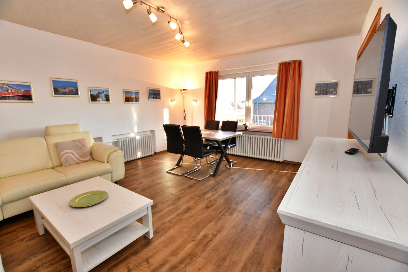 Wohn- und Essbereich mit Sofa, Esstisch und TV. Holzboden und Fenster mit Vorhängen.