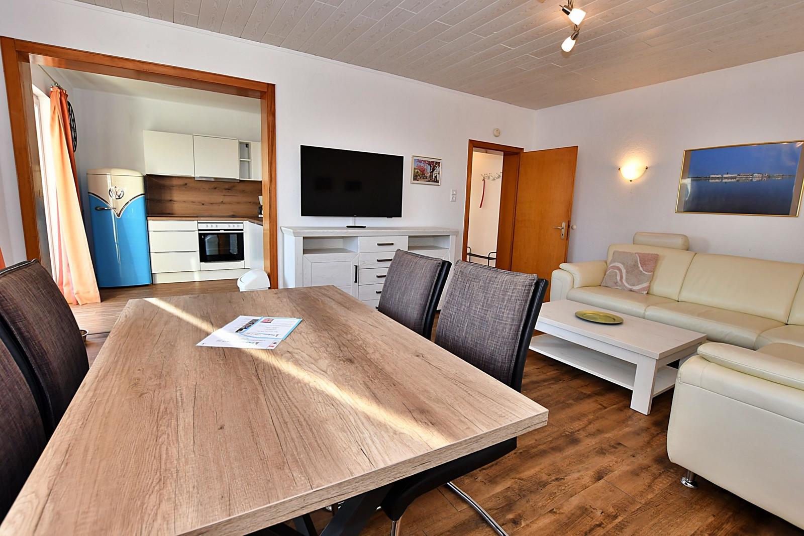 Essbereich mit Holztisch und Stühlen. Wohnzimmer mit Sofa, Couchtisch und Fernseher. Küche mit Kühlschrank und Herd.