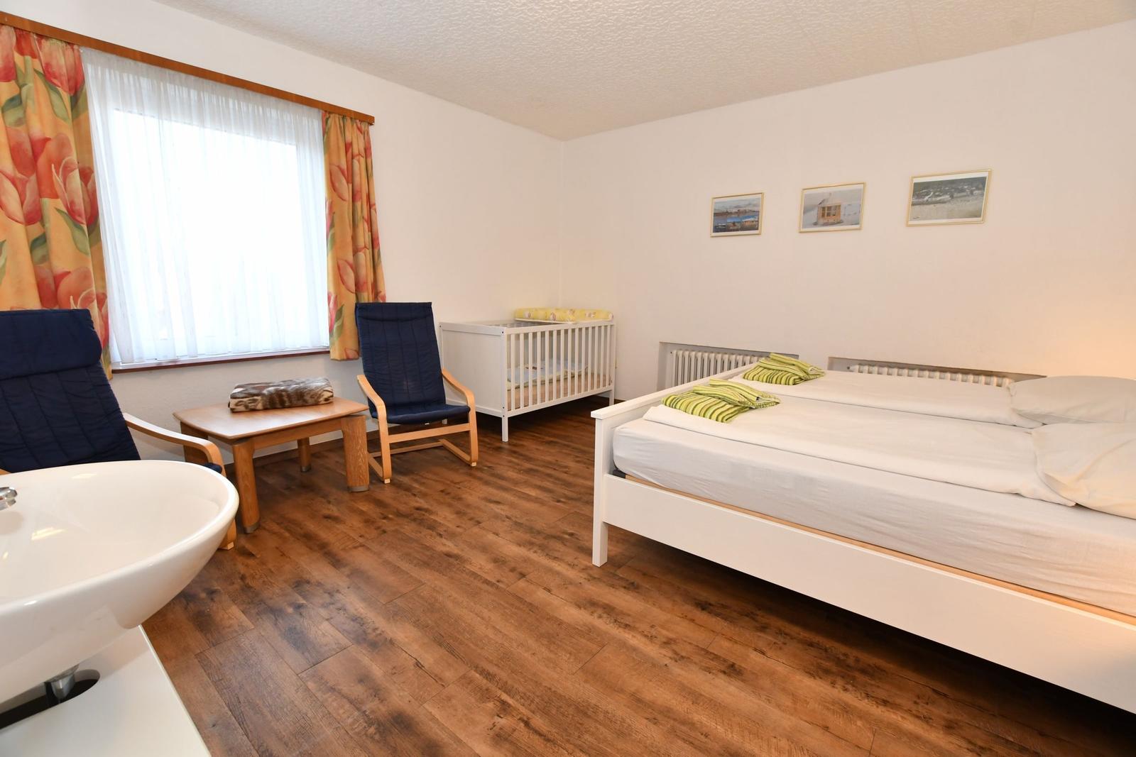 Zimmer mit Doppelbett, Kinderbett, zwei Stühlen und Fenster mit Blumenvorhängen.