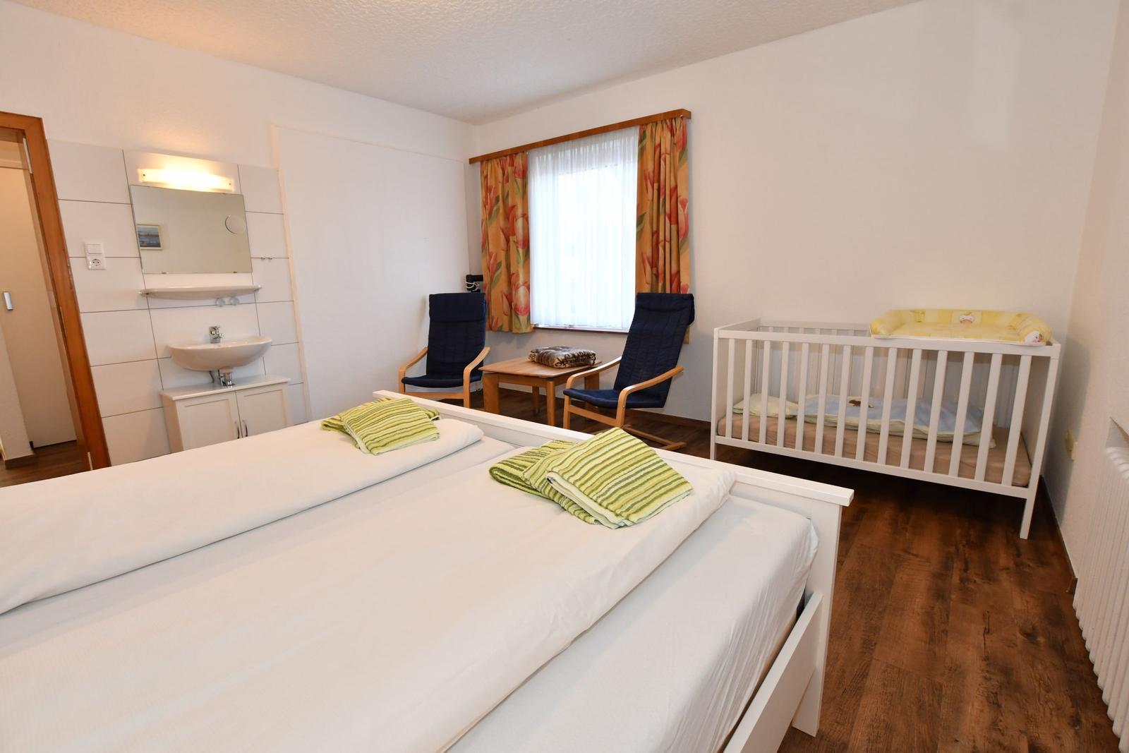Zimmer mit Doppelbett, Kinderbett, zwei Stühlen und Fenster mit Vorhängen.