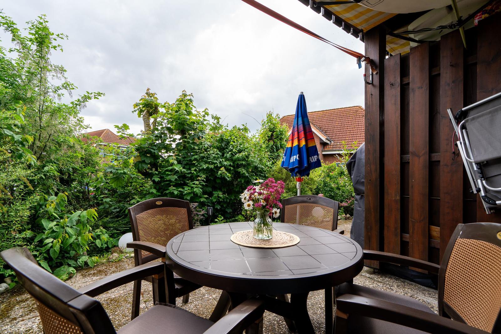 Round table with chairs and flowers on terrace with green surroundings.