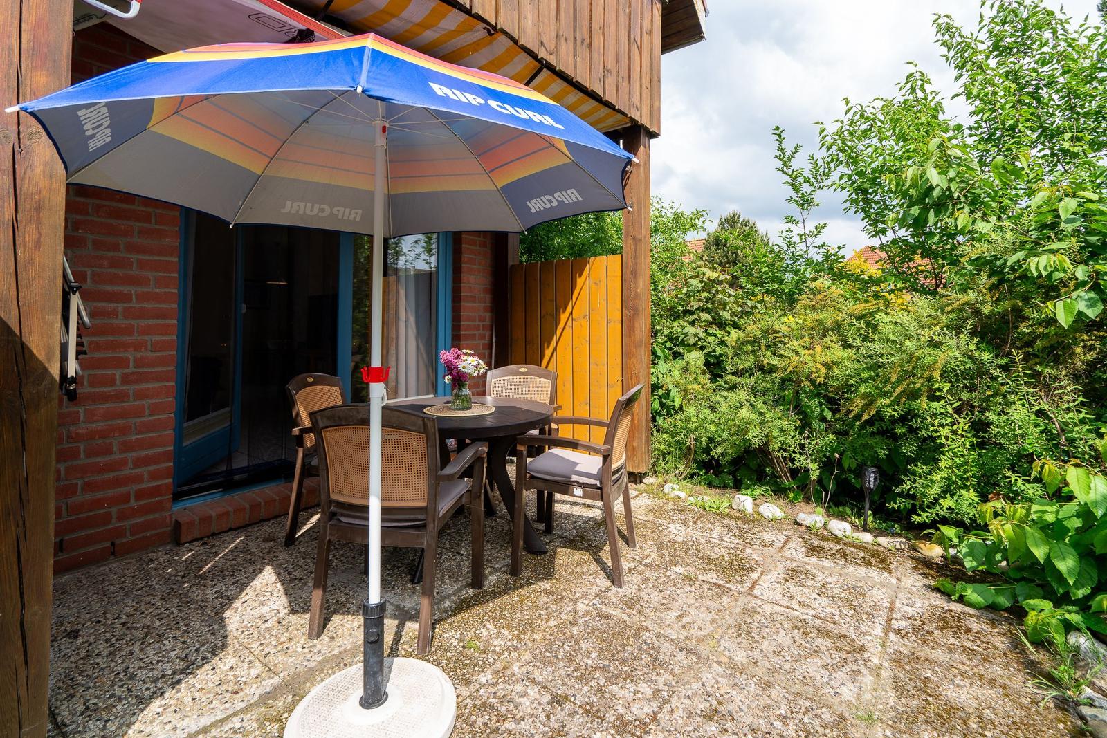 Terrace with table, chairs, and umbrella. Background: green plants and wooden house.