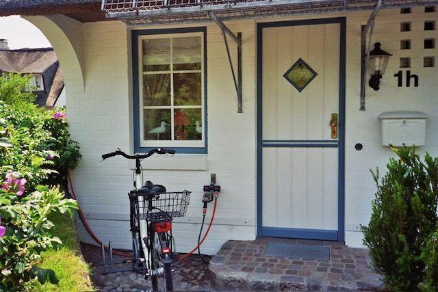 Eingangstür mit Fenster und Fahrrad vor einem weißen Haus mit Garten.