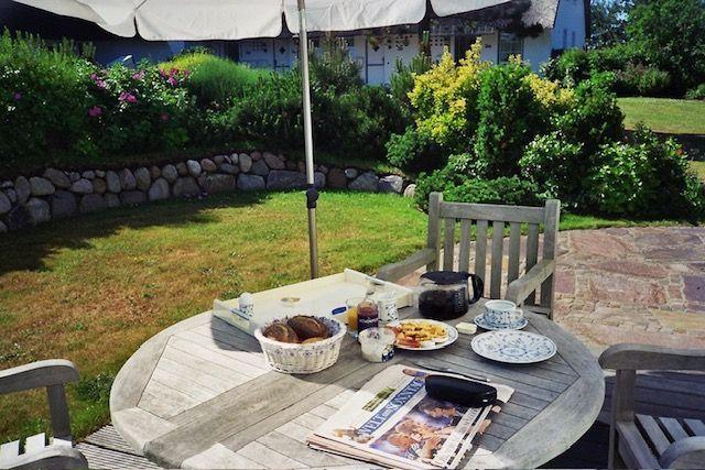 Außenbereich mit Tisch, Stühlen und Sonnenschirm. Auf dem Tisch stehen Frühstückszubehör und Zeitungen.