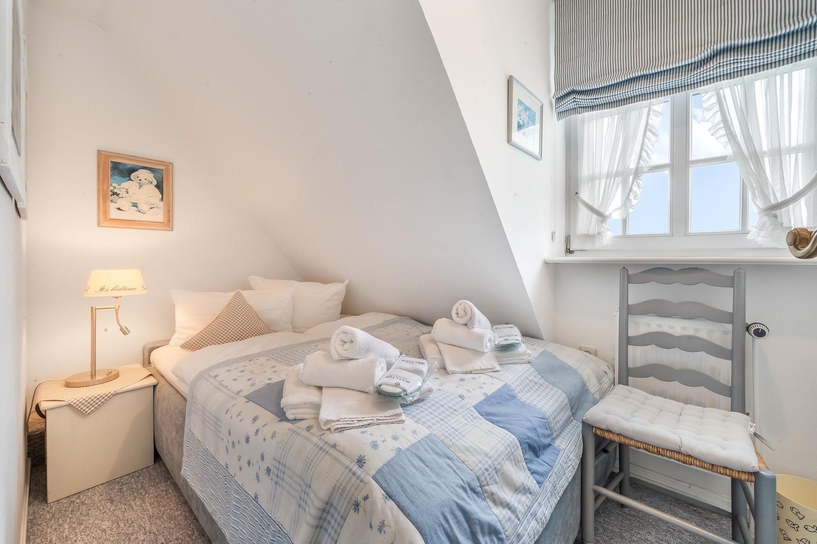 Schlafzimmer mit Dachschrägen, Bett mit Handtüchern, Fenster mit Vorhängen und Stuhl.