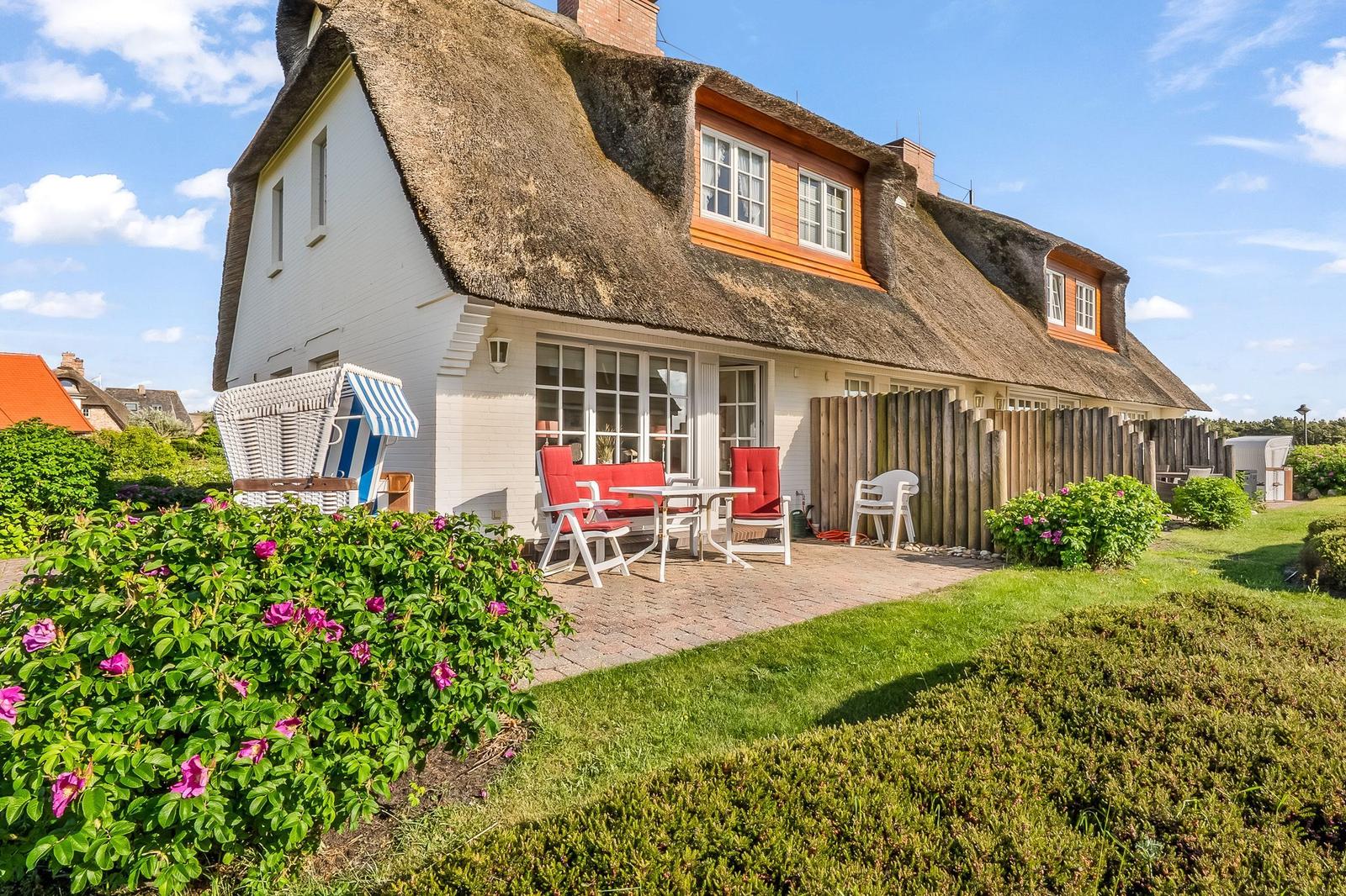 Haus mit Strohdach, Terrasse mit Tisch und Stühlen, Blumenbeete und Strandkorb.