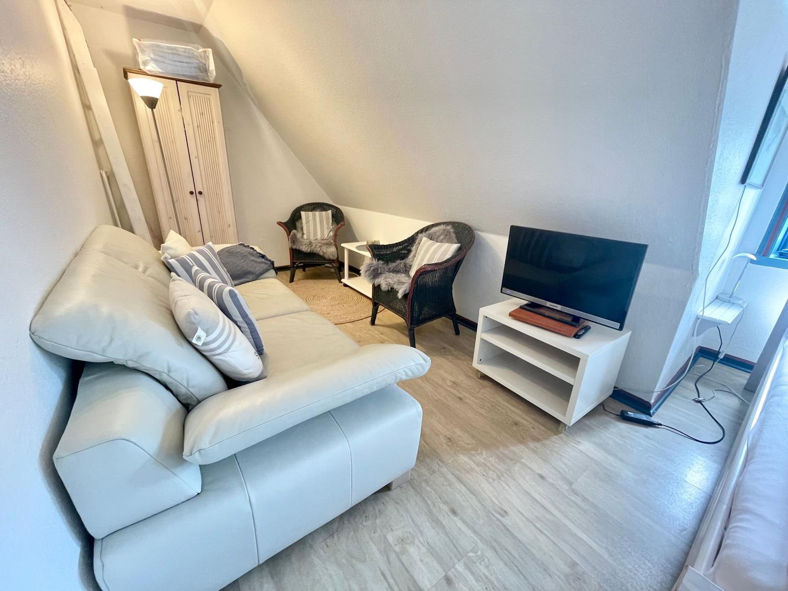 Wohnzimmer mit Sofa, zwei Stühlen, Fernseher und Kleiderschrank unter Dachschrägen.