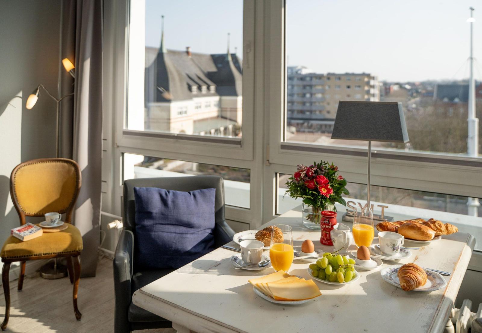 Frühstückstisch mit Obst, Brot und Getränken vor großem Fenster mit Stadtansicht.