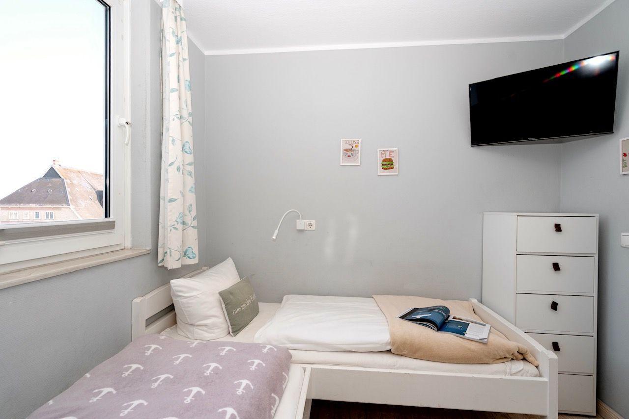 Ein Schlafzimmer mit Bett, Schrank und Fernseher an der Wand. Fenster mit Vorhang.