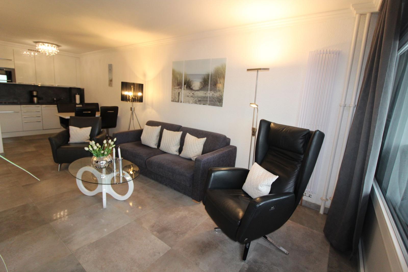 Living area with sofa, armchair, and glass table. Kitchen in background.