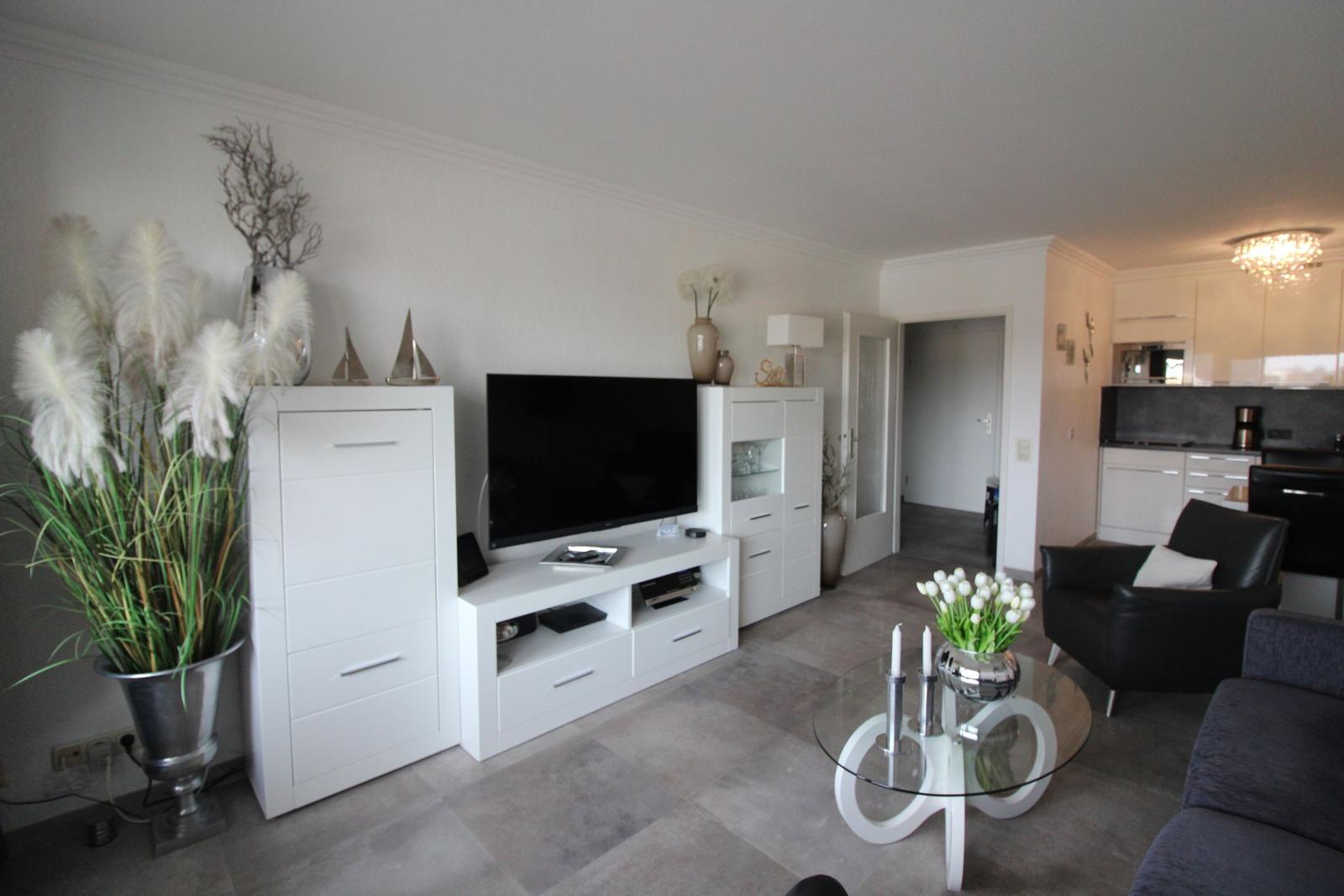 Living room with TV, glass table, and seating. Kitchen in the background.
