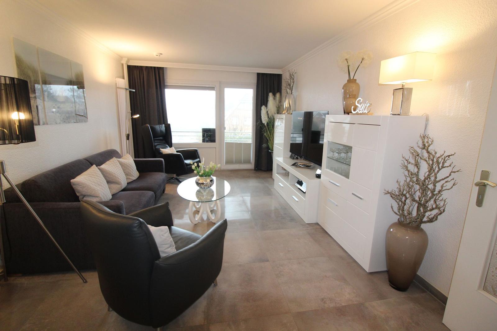 Living room with sofa, armchairs, glass table, and TV. Terrace with view outside.